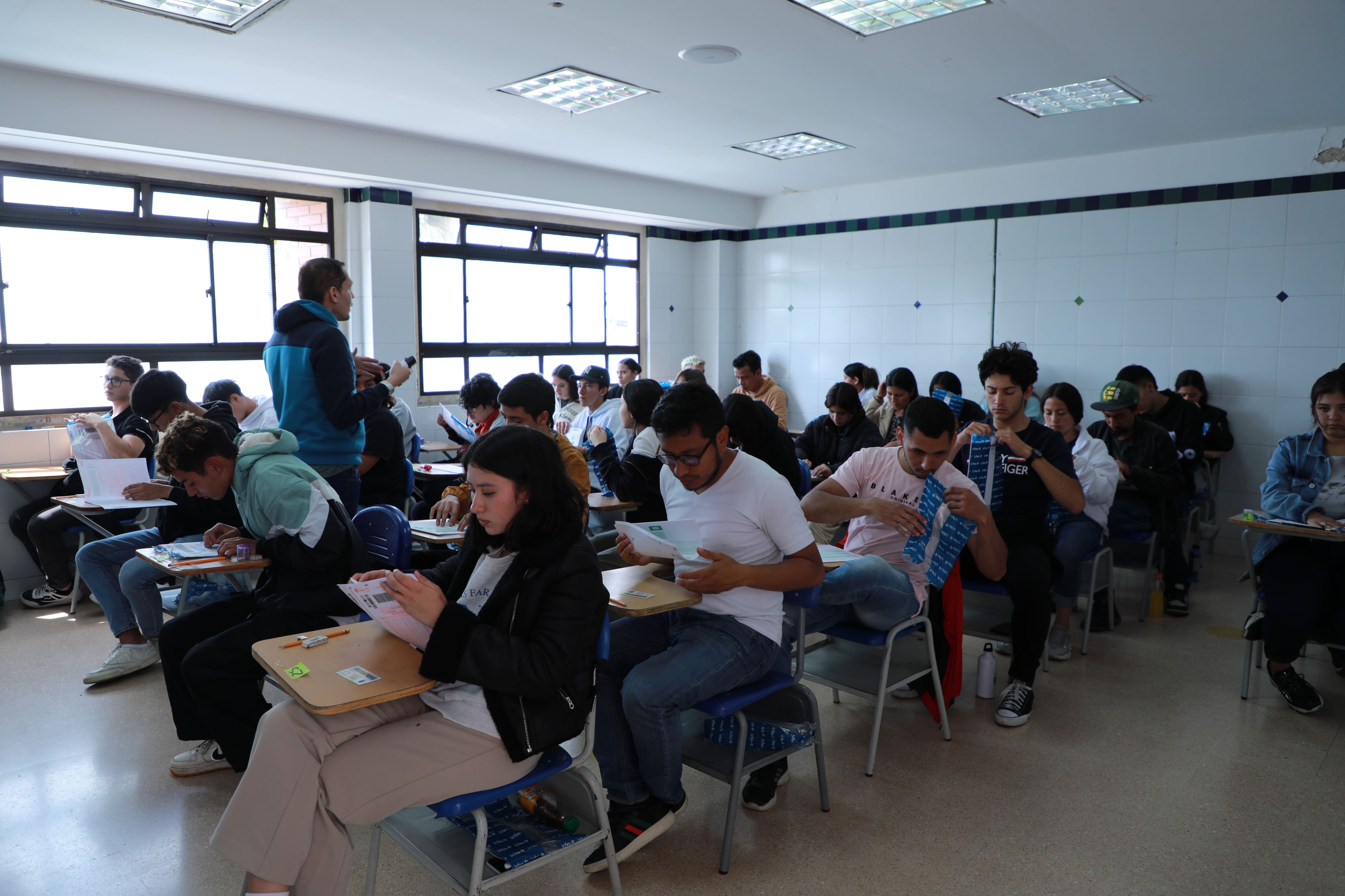 Jóvenes en salón de clase con profesor que da instrucciones mientras ellos están por responder examen del ICFES